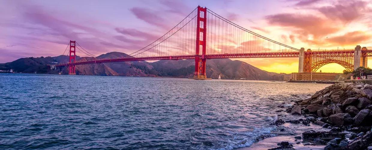 Il Golden Gate Bridge al tramonto con un cielo multicolore e la Baia di San Francisco in primo piano.