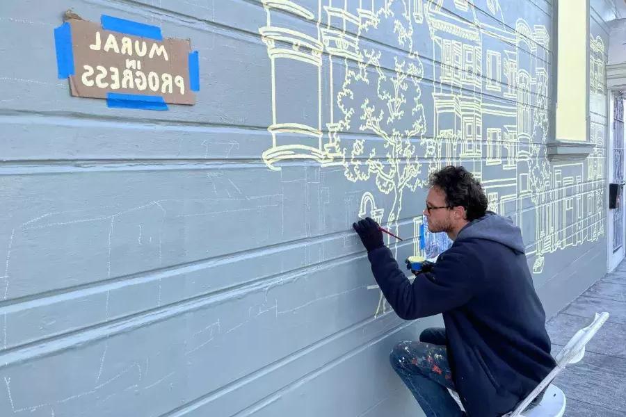 一位艺术家在教会区一栋建筑的侧面画壁画, 在建筑上贴了一个牌子，上面写着“壁画正在进行中”." San Francisco, CA.